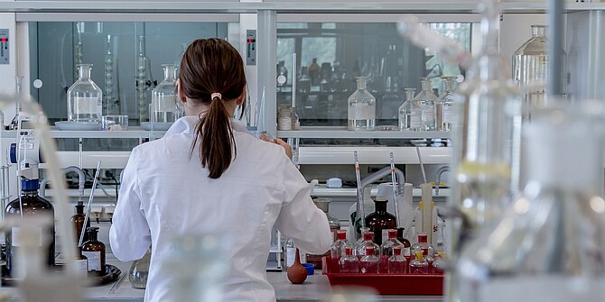 Eine Forscherin arbeitet im Labor. Sie ist von hinten aufgenommen. Um sie herum sieht man Glasfläschchen und Laborausrüstung.