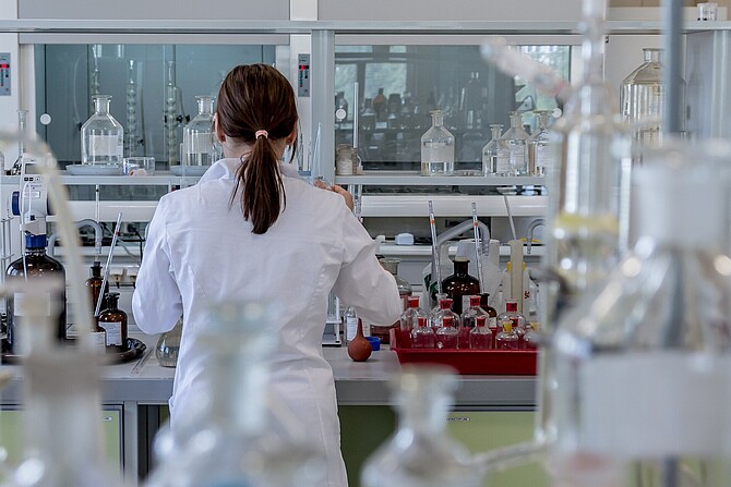 Eine Forscherin arbeitet im Labor. Sie ist von hinten aufgenommen. Um sie herum sieht man Glasfläschchen und Laborausrüstung.